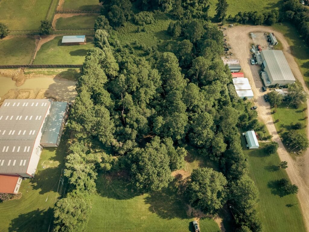 Aerial view of a wooded lot before land clearing, dense tree canopy running the length of the property between a metal barn and a shed row, near Navasota, TX