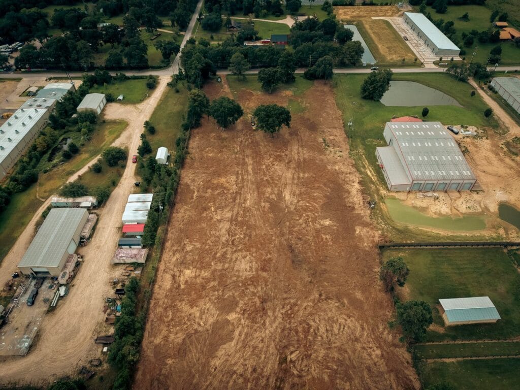 The same end of the lot after clearing, a wide strip of open graded ground with a mature oak left standing and an excavator working, near Navasota, TX